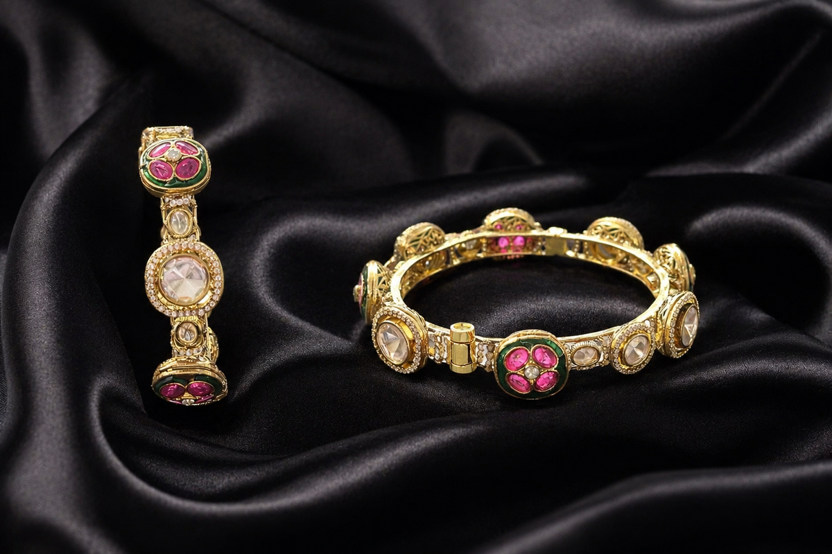 A gold Kundan-style bangle and matching ring set with clear uncut stones and pink and green enamel floral accents, displayed on draped black satin fabric.