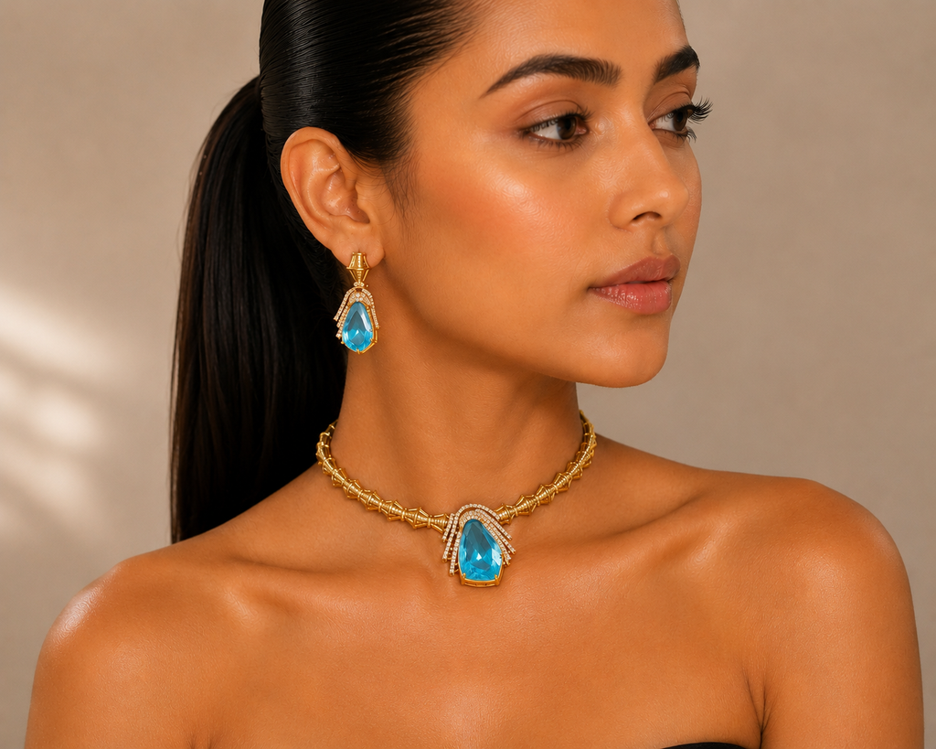 A woman with dark hair in a ponytail models a gold jewelry set featuring a braided chain necklace and matching drop earrings, each with a teardrop-shaped light blue gemstone.