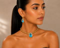 A woman with dark hair in a ponytail models a gold jewelry set featuring a braided chain necklace and matching drop earrings, each with a teardrop-shaped light blue gemstone.