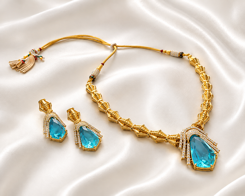 A gold necklace and earring set with pear-shaped light blue gemstones bordered by pavé stones and a braided tasselled cord closure, displayed on draped white satin fabric.