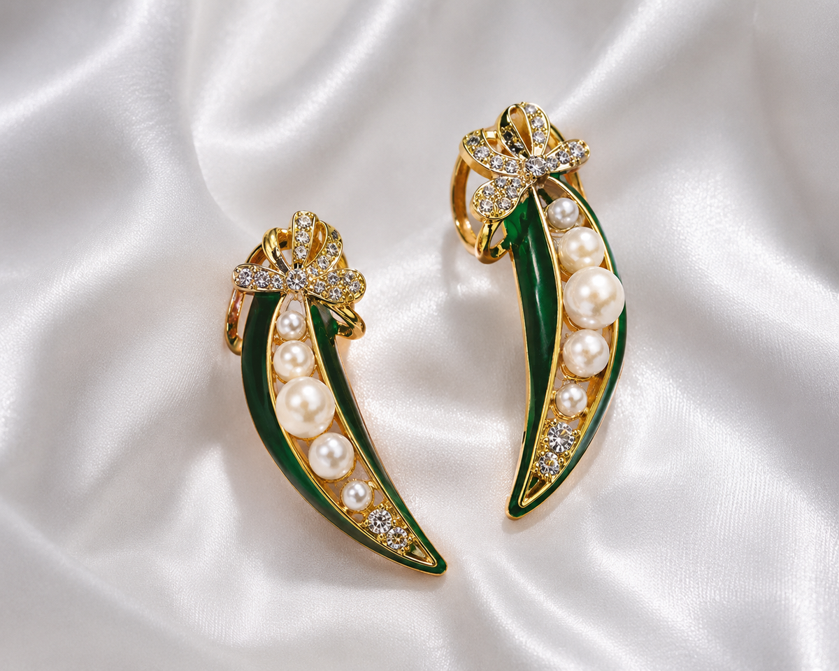 A pair of gold and green enamel leaf-shaped earrings with white pearls and clear rhinestone accents, displayed on a white satin fabric background.