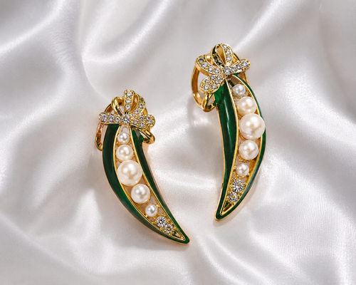 A pair of gold and green enamel leaf-shaped earrings with white pearls and clear rhinestone accents, displayed on a white satin fabric background.