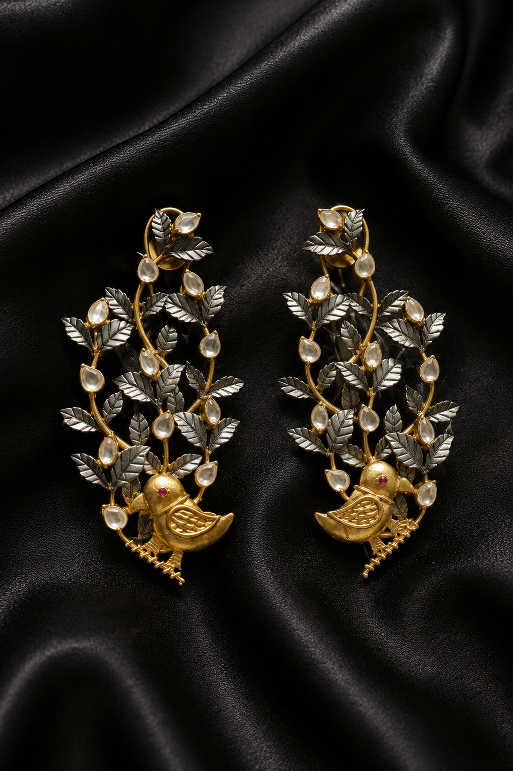 A pair of two-tone gold and silver bird-and-leaf motif drop earrings with pearl accents, displayed on draped black satin fabric.