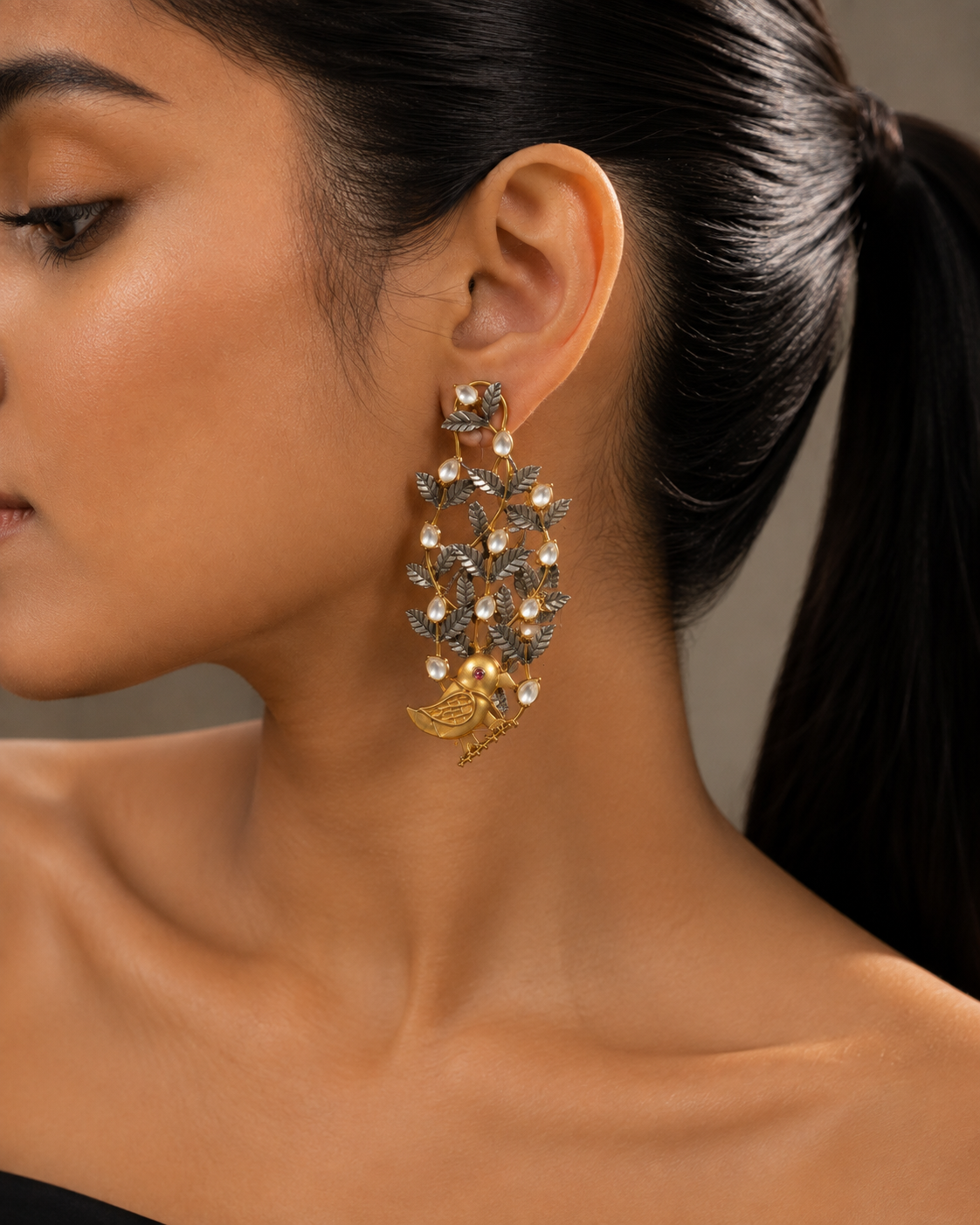 A close-up side profile of a model wearing a large two-tone gold and silver bird-and-leaf motif dangle earring with pearl accents.