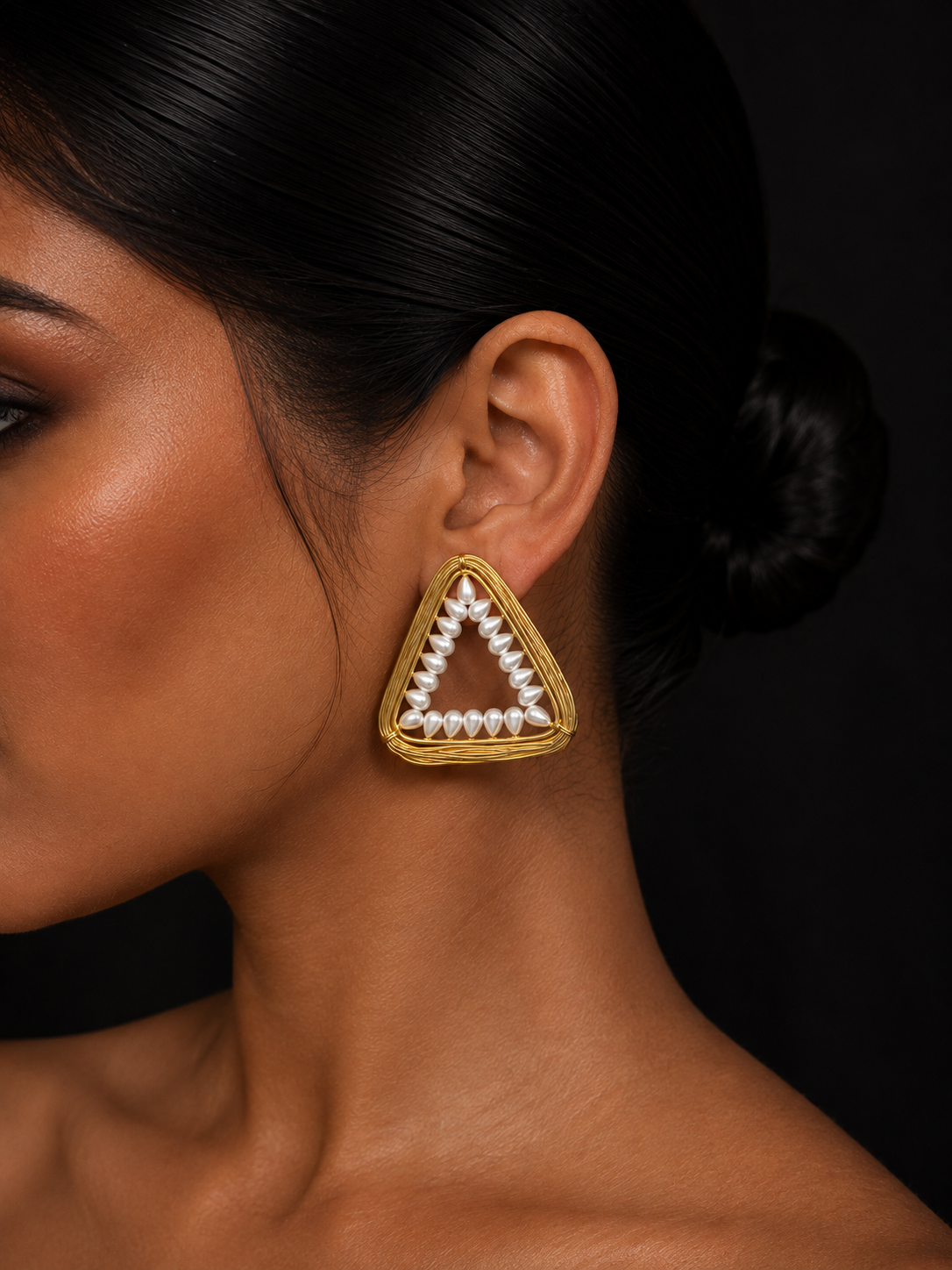 A close-up of a woman's ear and neck wearing a gold triangular stud earring with a white pearl border, set against a dark background.