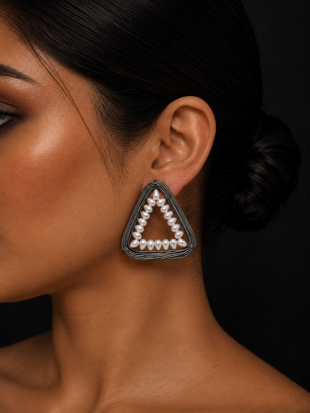 A close-up of a woman's ear and side profile wearing a triangular statement earring with a black textured frame and a border of white faux pearls, set against a dark background.