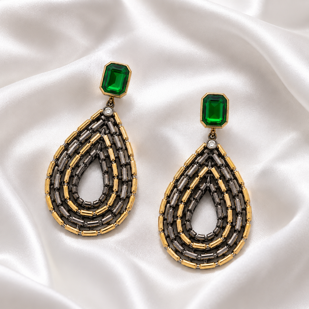 A pair of large teardrop-shaped gold and gunmetal beaded earrings with emerald-green rectangular stones, displayed on draped white satin fabric.