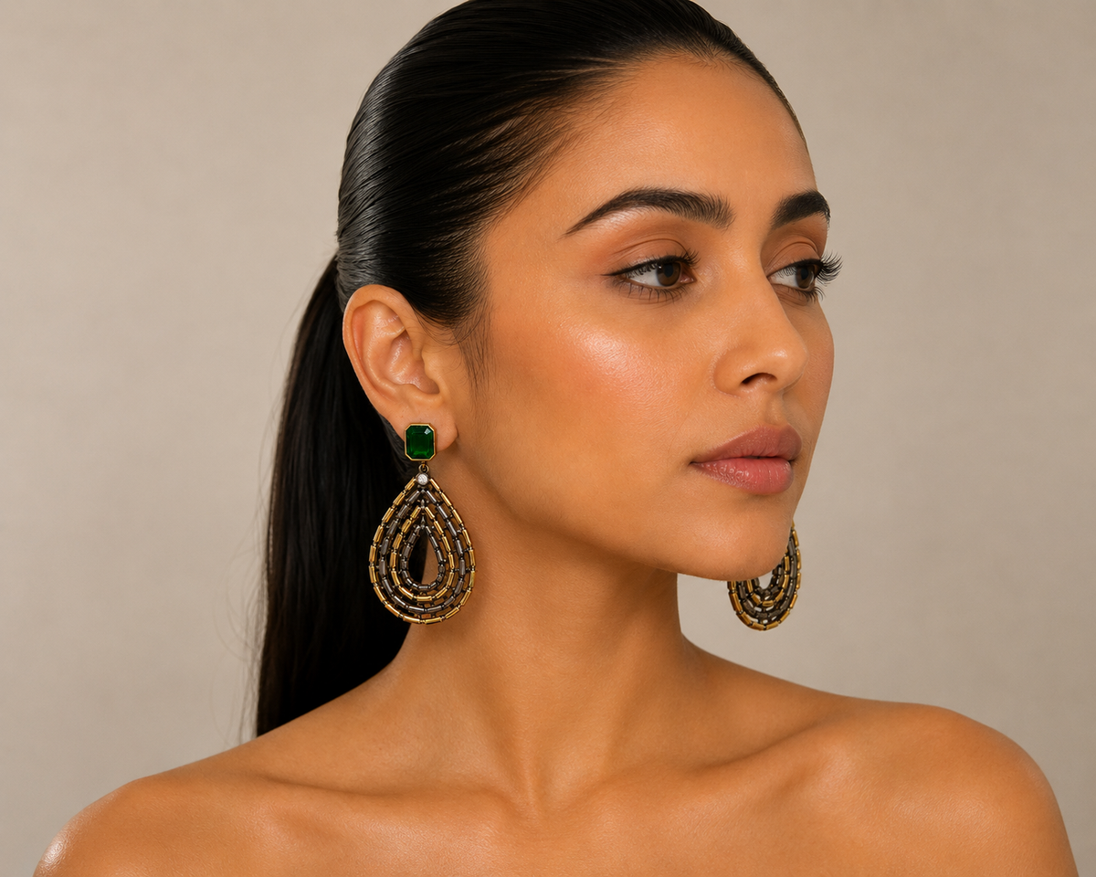 A woman with dark hair in a ponytail wearing large gold teardrop earrings with green square accents, shown in a close-up portrait against a neutral beige background.