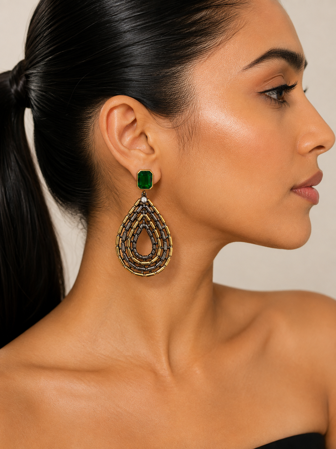 A close-up side profile of a woman wearing a large teardrop-shaped gold dangle earring with a green rectangular stone and intricate gold detailing.