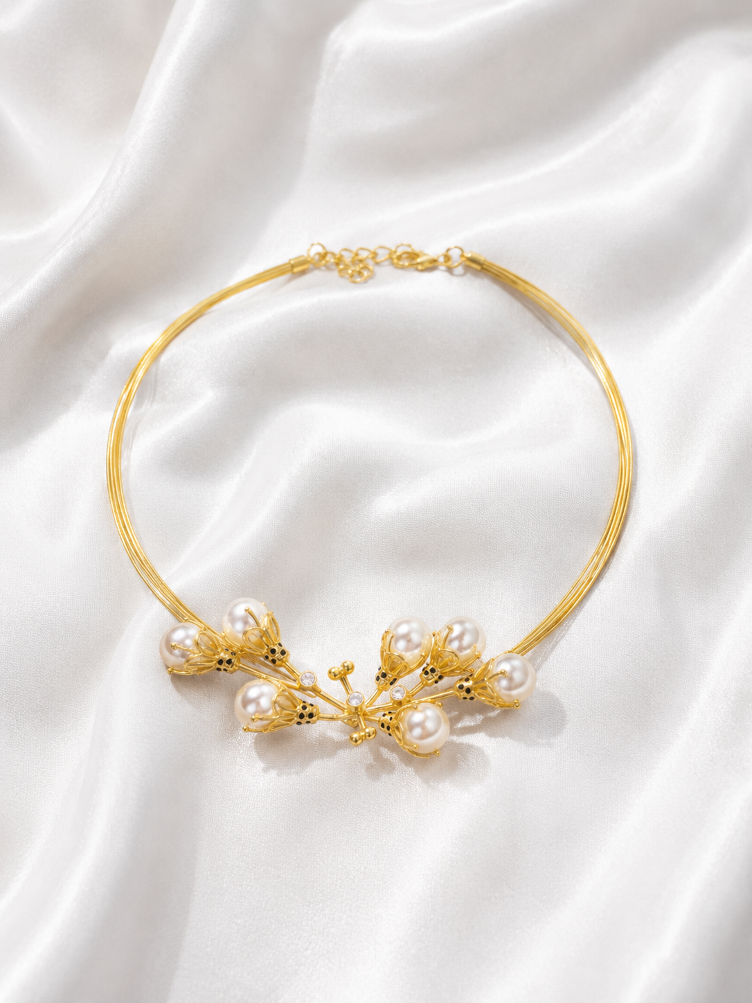 A delicate gold wire choker necklace featuring a central cluster of white pearls and small gold floral details, displayed on a draped white satin fabric background.