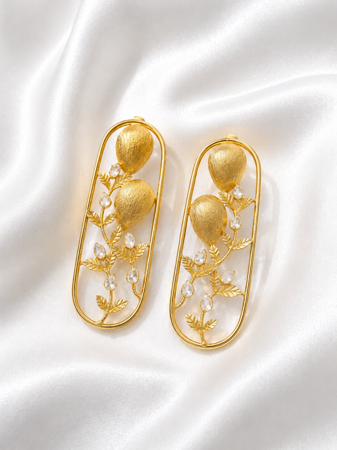 A pair of gold-toned statement earrings with textured oval frames, leafy vine details, and clear faceted crystal accents, displayed on a white satin fabric background.