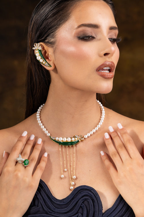 A close-up of a woman wearing a pearl and green enamel necklace with dangling chains, matching pearl ear cuff, and a pearl ring with a green stone, against a dark brown background.