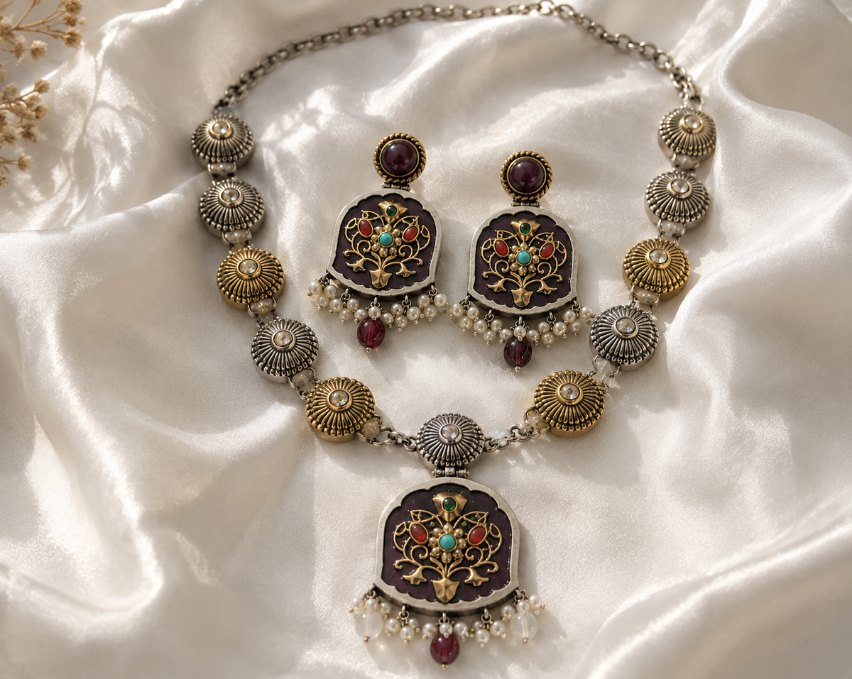 An ornate jewelry set featuring a necklace and matching earrings with dark purple enamel, gold filigree, and red and turquoise accents, displayed on a draped white satin fabric.