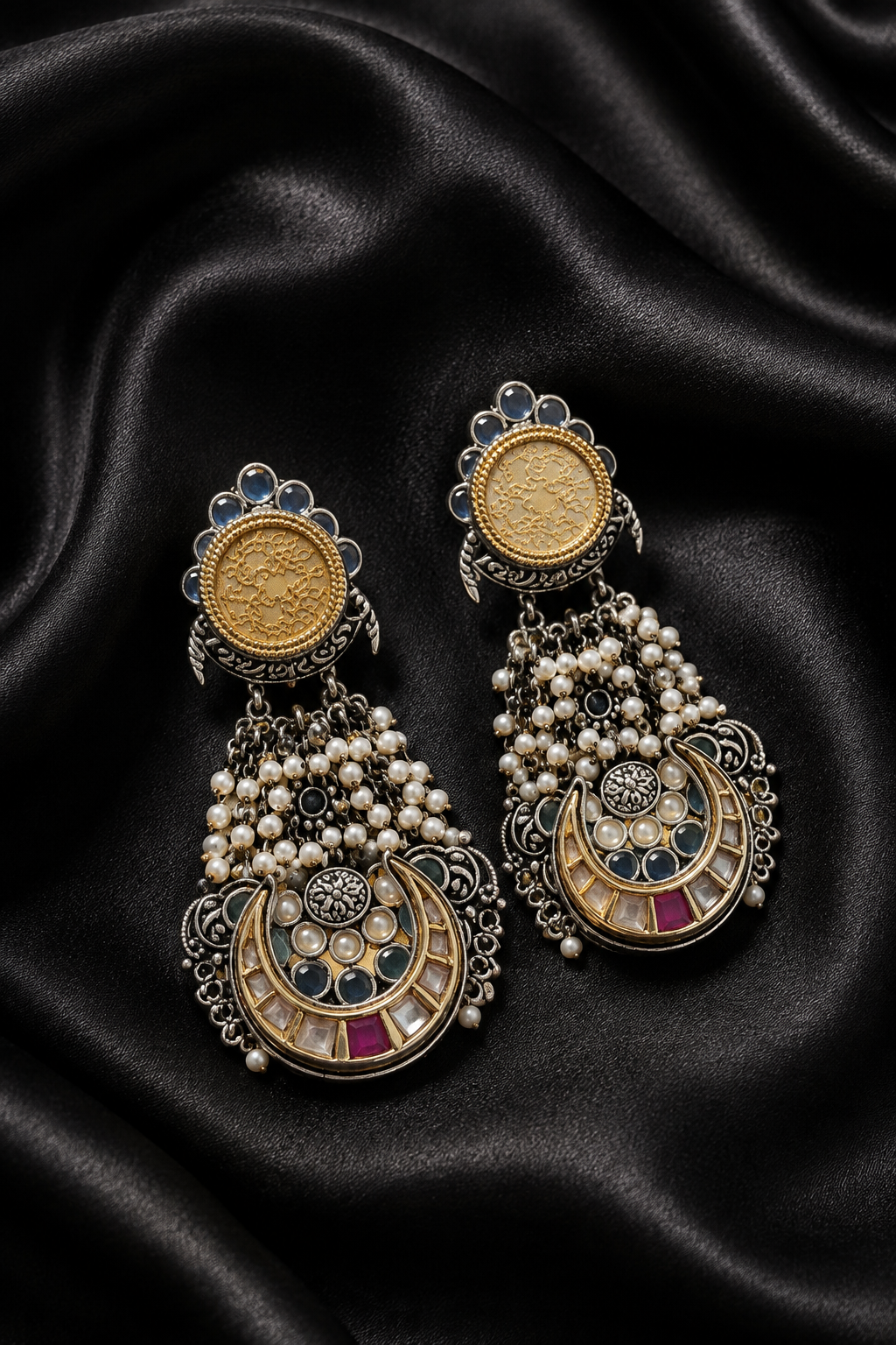 A pair of ornate gold-tone crescent-shaped drop earrings with pearl clusters and blue and pink stone accents, displayed on draped black satin fabric.
