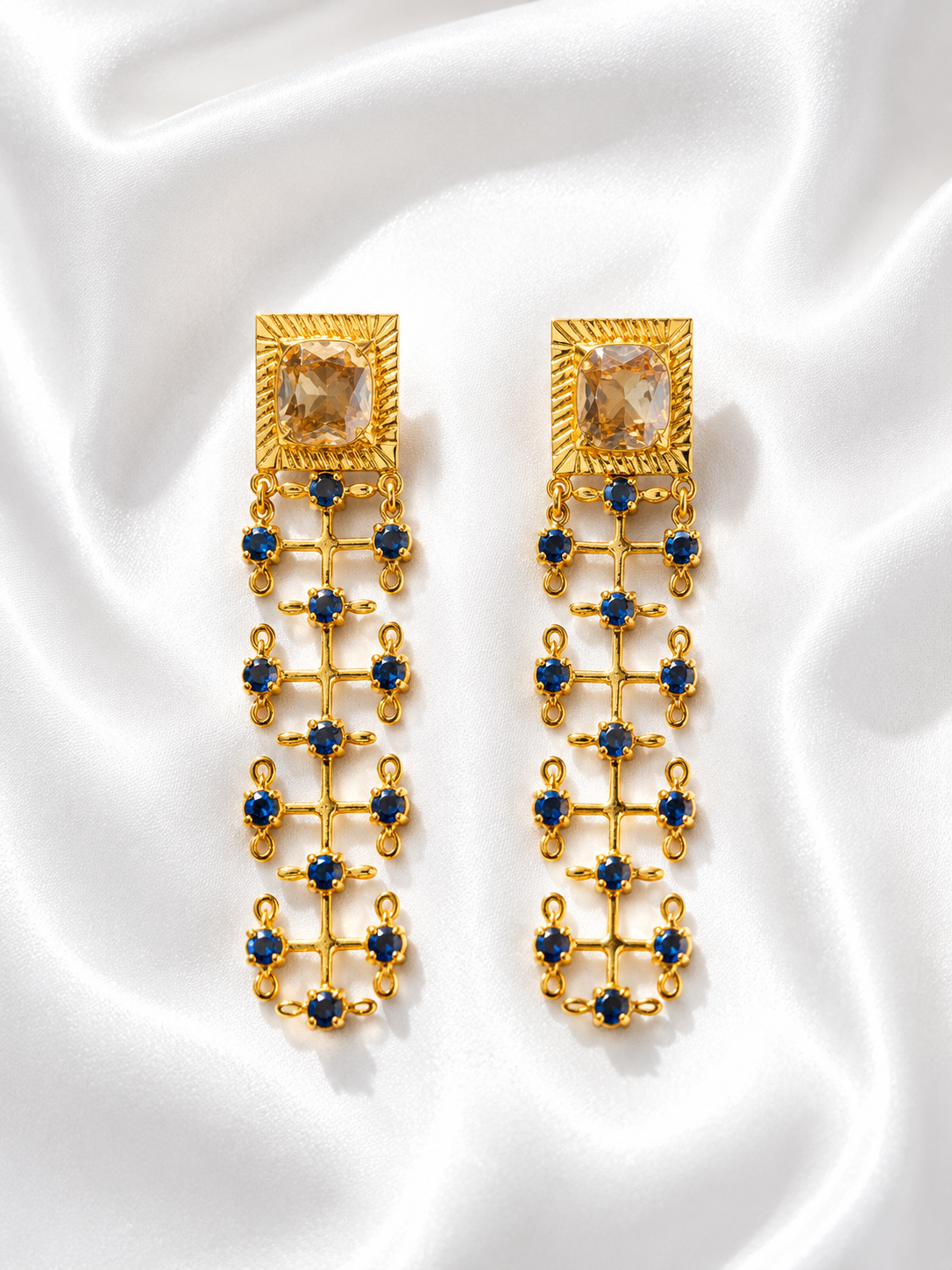 A pair of gold dangle earrings with a large faceted clear crystal at the top, accented by rows of small round blue gemstones, displayed on a draped white satin fabric.