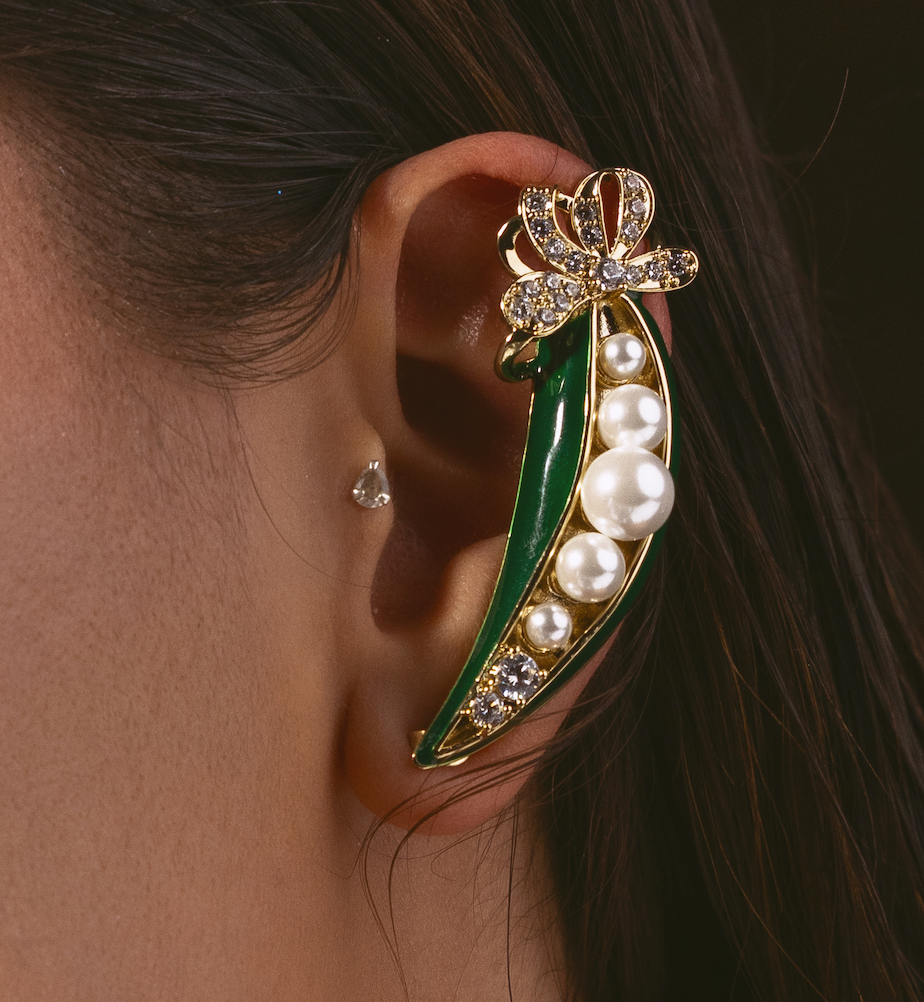 A close-up of a model's ear wearing a gold ear cuff designed like a green pea pod with white pearls and a rhinestone-embellished bow at the top.