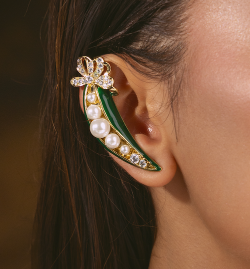 A close-up of a model's ear wearing a green pea pod ear cuff. The ear cuff features a gold-tone metal frame, green enamel, white faux pearls, and clear rhinestones forming a bow at the top.