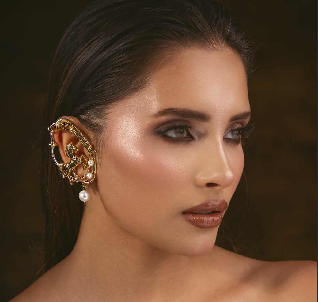 A close-up portrait of a woman wearing an ornate gold ear cuff with small white pearl accents, set against a dark brown background.
