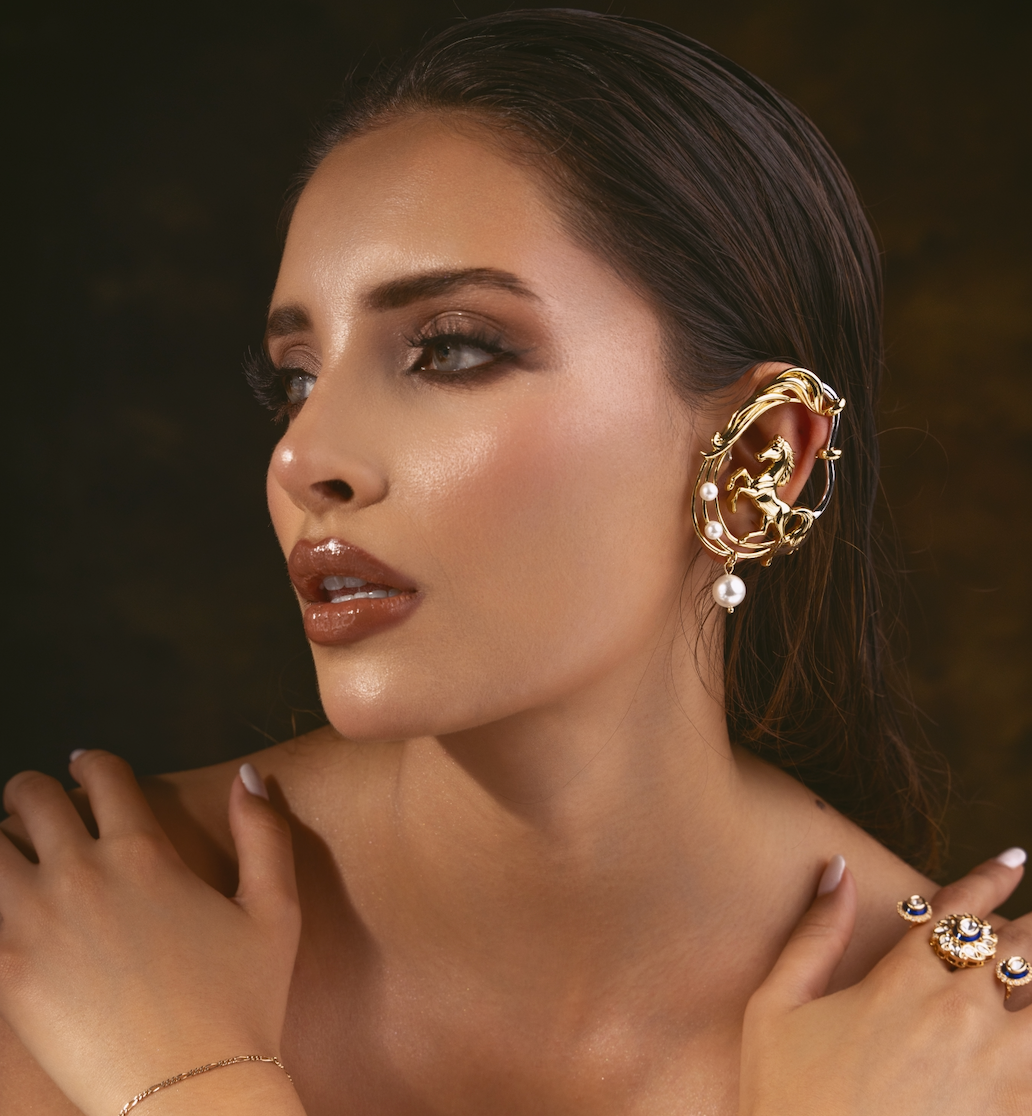 A close-up portrait of a woman wearing an ornate gold ear cuff with dangling pearl-like accents, along with rings and a delicate bracelet, against a dark warm-toned background.