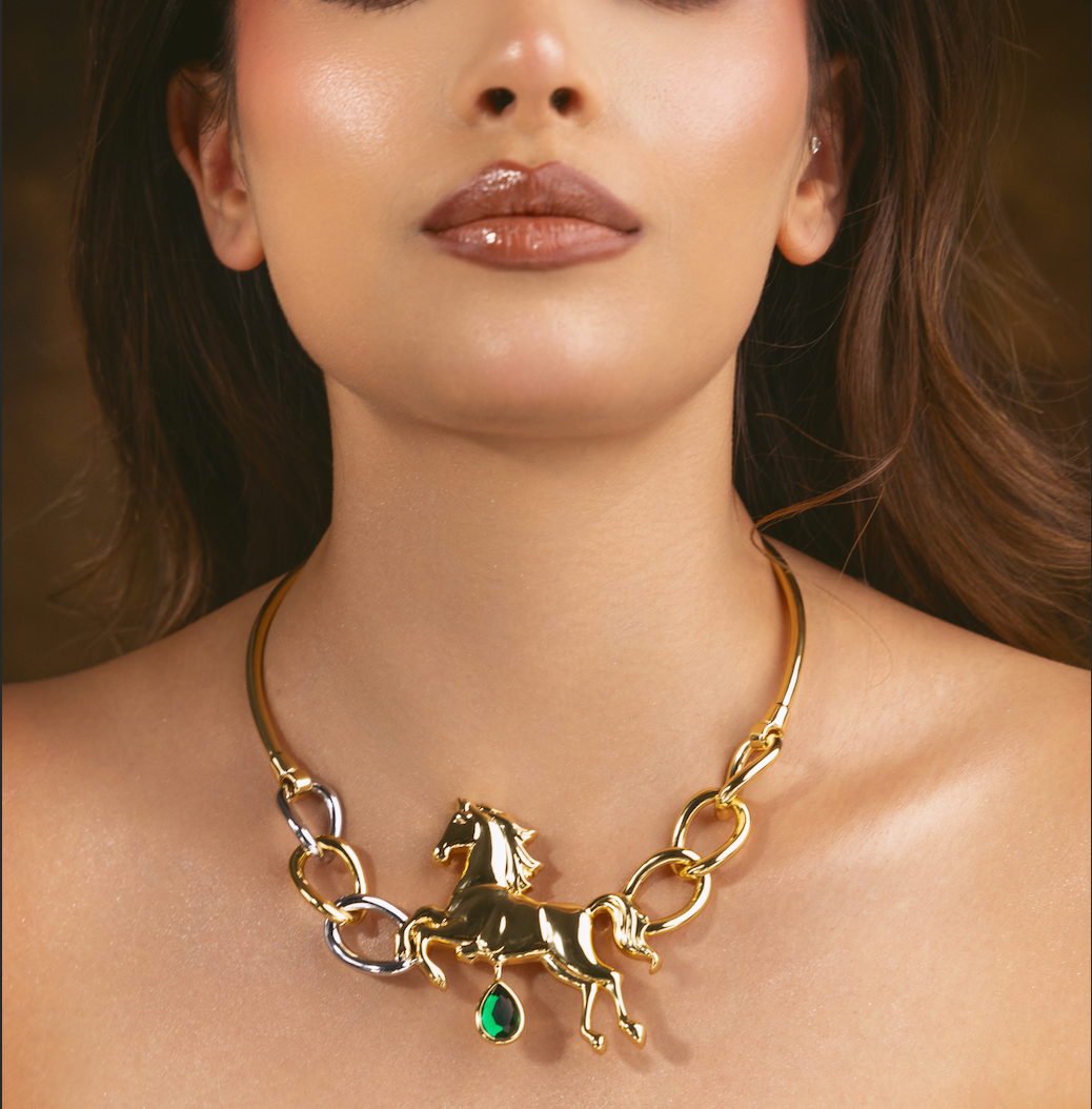 A close-up of a model wearing a gold chain necklace with a horse charm and a green teardrop accent, set against a warm, blurred background.