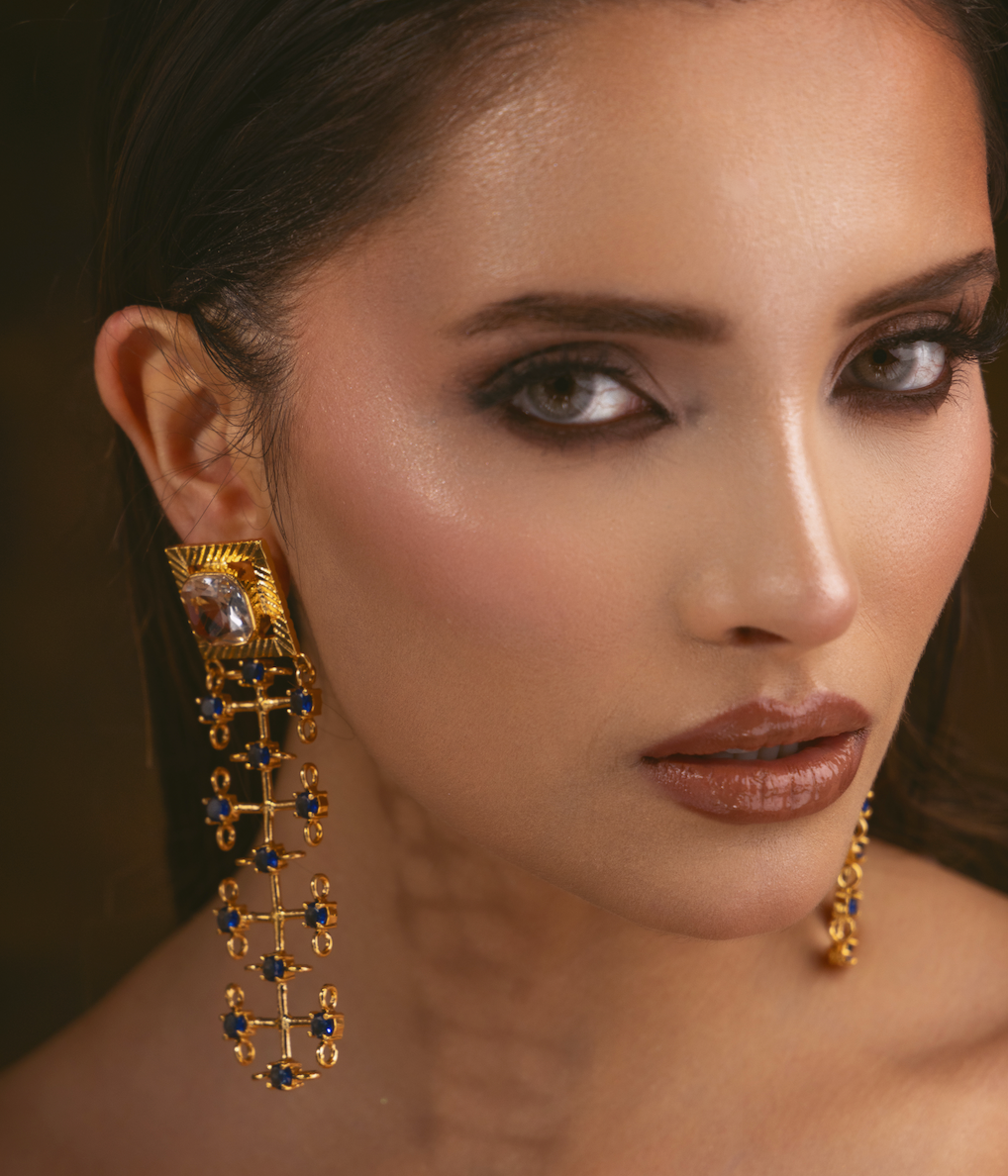 A close-up portrait of a woman wearing a dramatic gold statement earring with blue gemstone accents and a large clear central stone, against a dark blurred background.