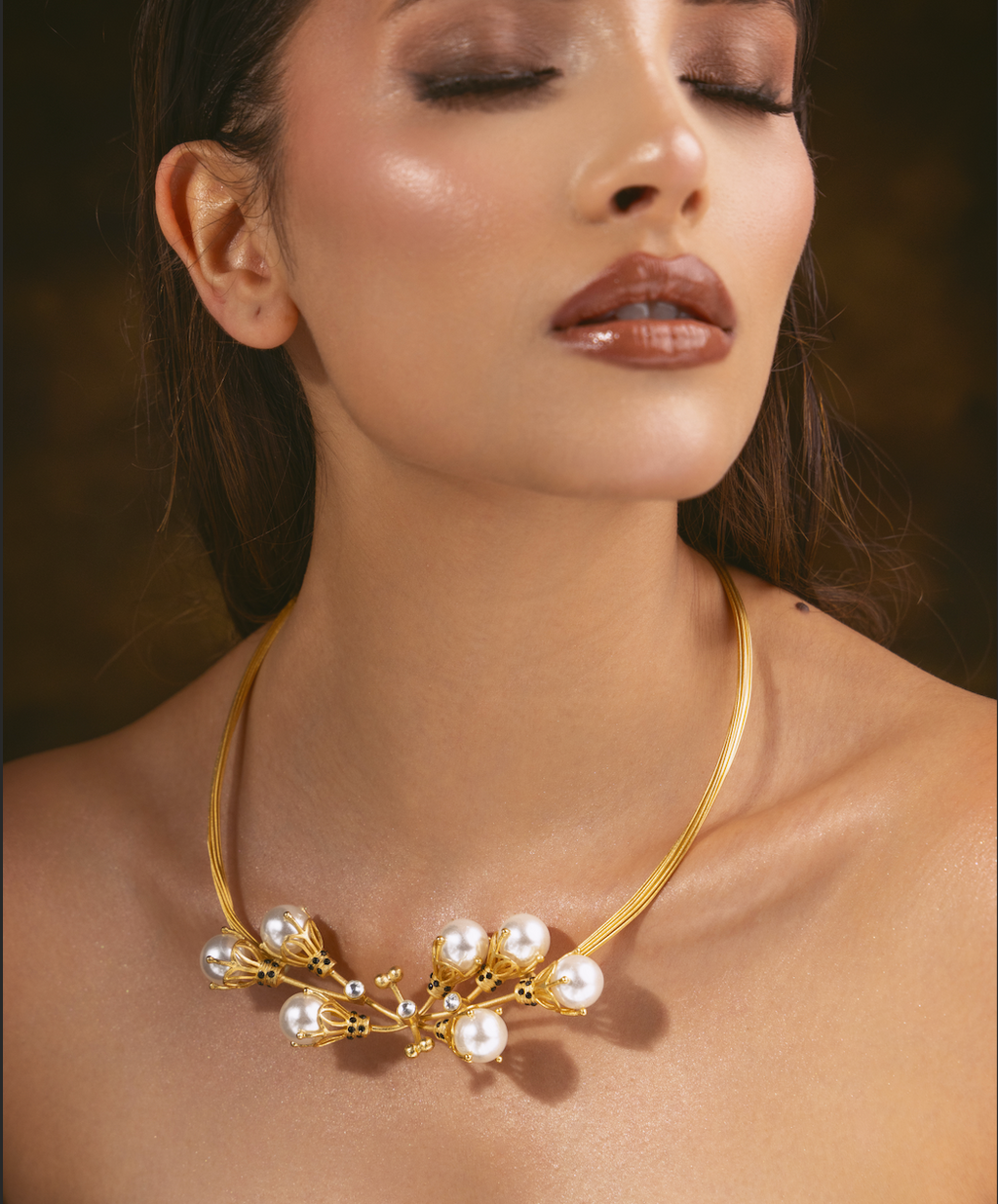 A close-up of a woman wearing a gold necklace with white pearls and small gold floral details, set against a dark brown background.