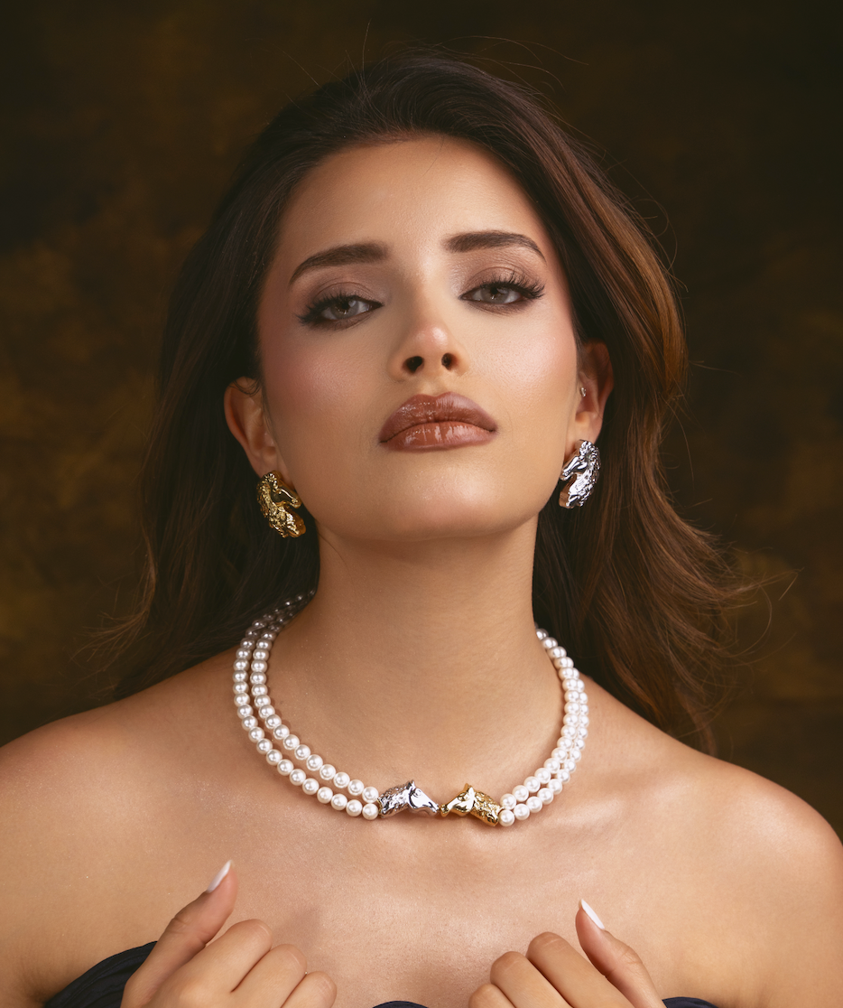 A close-up portrait of a woman wearing a double-strand pearl necklace with horse-shaped silver and gold pendants, matching horse earrings, and a dark strapless top against a warm brown background.