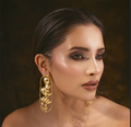 A close-up portrait of a woman wearing a large gold statement earring with sculptural floral elements and clear rhinestone accents, set against a dark brown background.