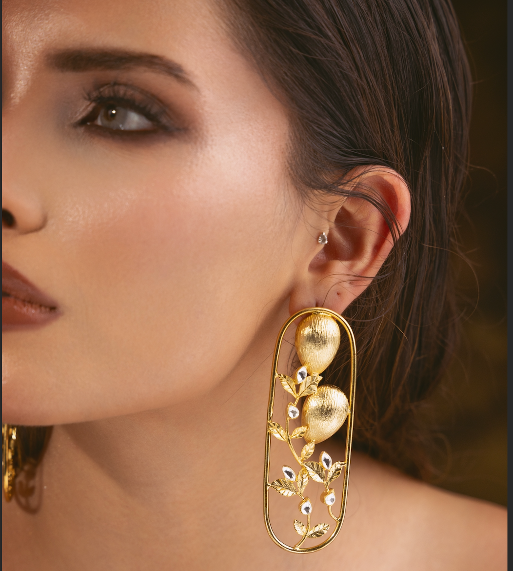 A close-up of a woman's ear wearing a large gold statement earring with elongated teardrop-shaped gold elements and small white stone accents, against a dark warm-toned background.