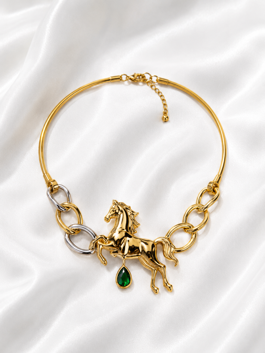 A gold-toned bangle necklace featuring a detailed horse charm, silver-toned chain links, and a dangling green teardrop gemstone, displayed on a white satin fabric background.