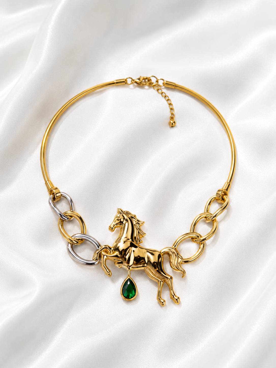 A gold-toned bangle necklace featuring a detailed horse charm, silver-toned chain links, and a dangling green teardrop gemstone, displayed on a white satin fabric background.