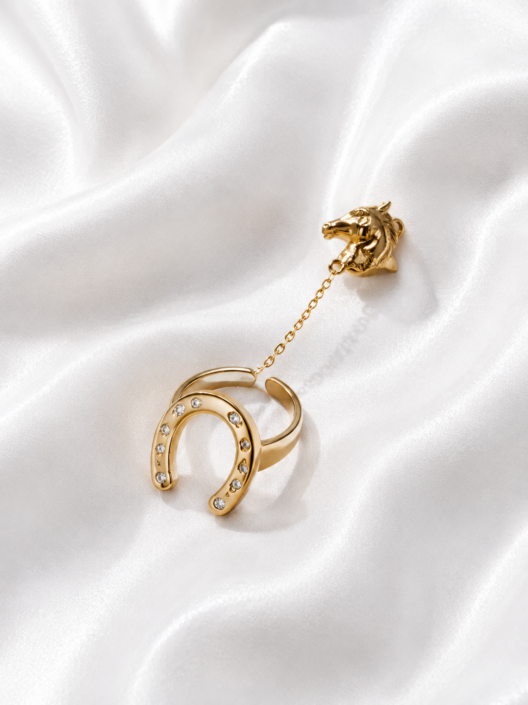A gold-tone ring with a horse head charm connected by a delicate chain to a horseshoe-shaped band set with small clear stones, displayed on draped white satin fabric.